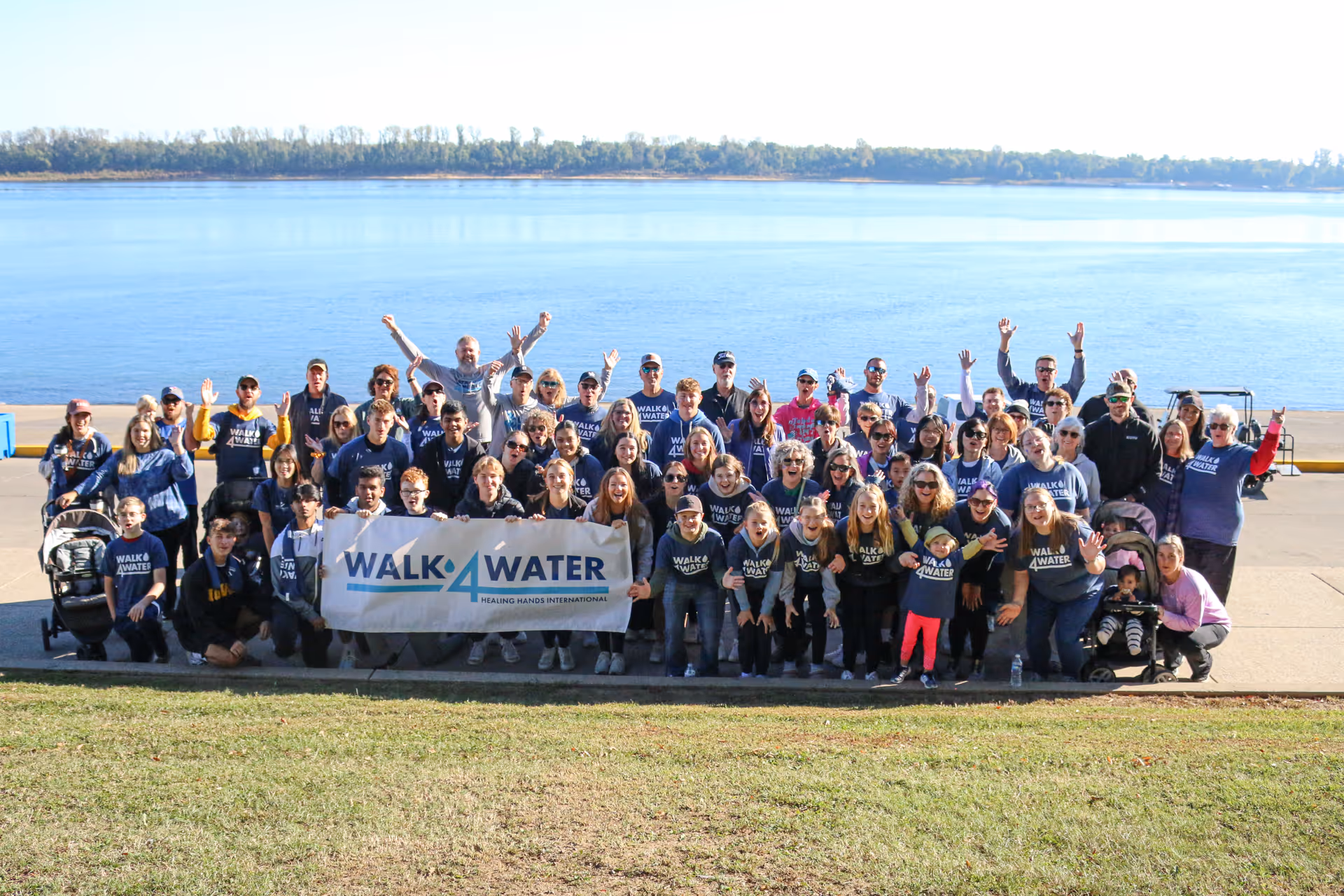 Large group of people outdoors by a river holding a Walk4Water banner, many wearing matching Walk4Water shirts and waving.