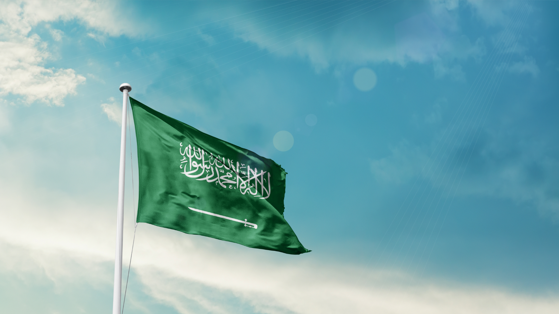 Saudi Arabian flag with white Arabic script and sword on green, waving against a blue sky.