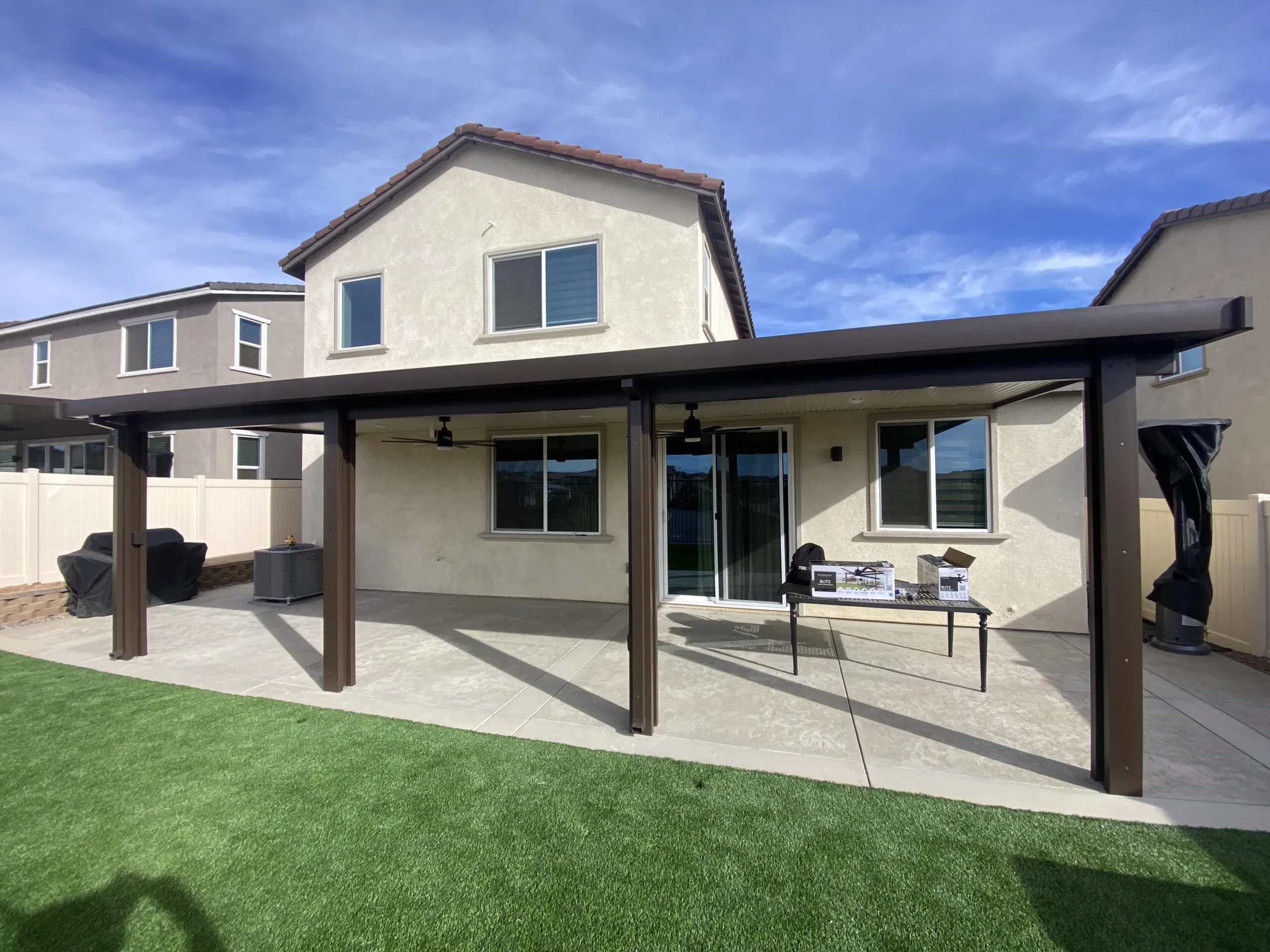 Backyard of a two-story house with a covered patio, ceiling fans, and a table holding boxes next to sliding glass doors.