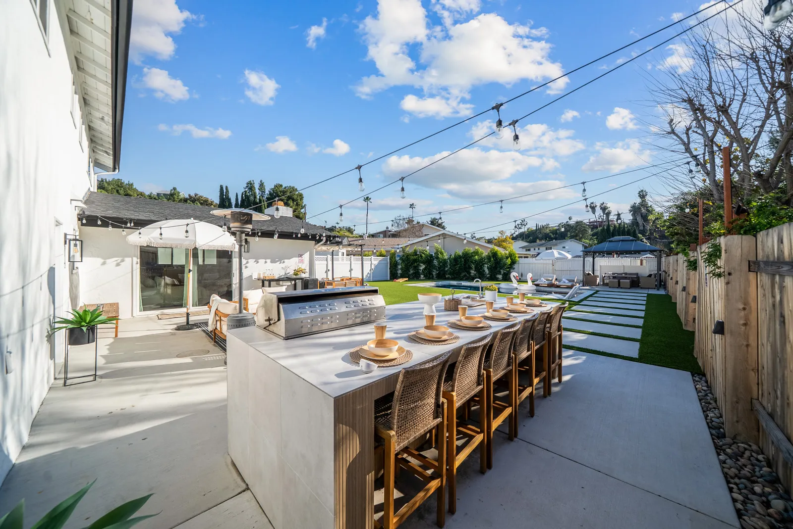 Outdoor patio with a long dining table set with plates and cups, bar stools, a built-in grill, string lights overhead, lounge chairs, and a pool with inflatable swans under a partly cloudy sky.