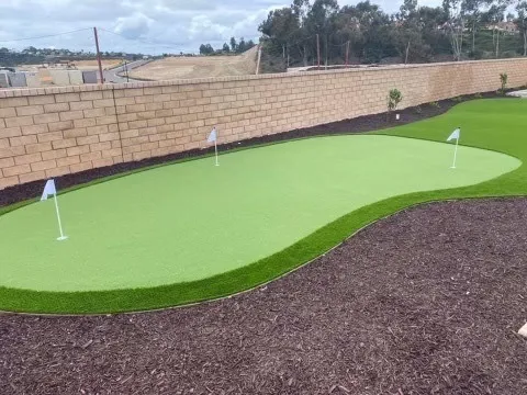 Artificial putting green with three golf holes and white flags in a backyard next to a brick wall.