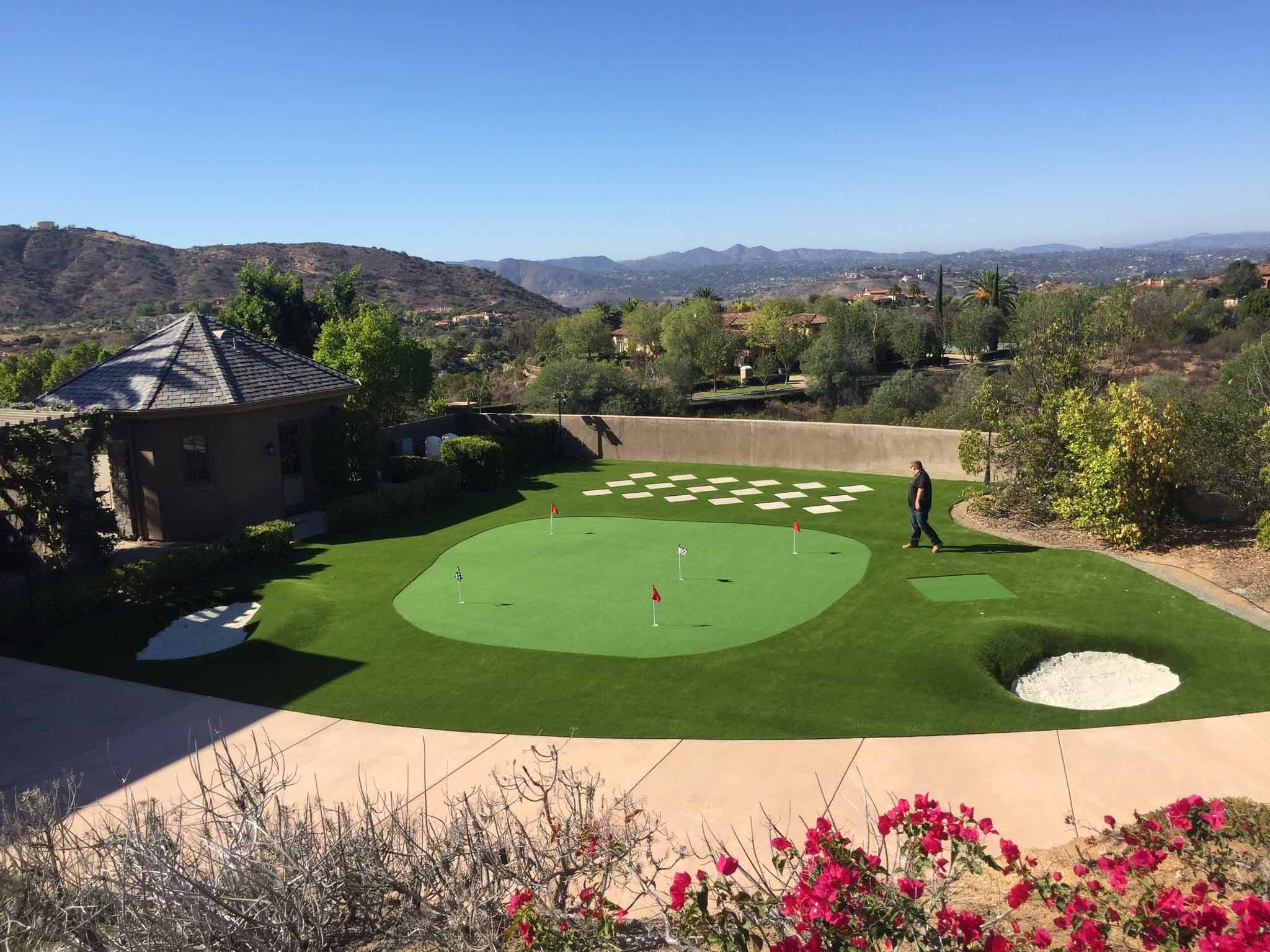 Backyard patio with a covered pergola, ceiling fans, a table holding boxes, artificial grass, and a two-story beige house.