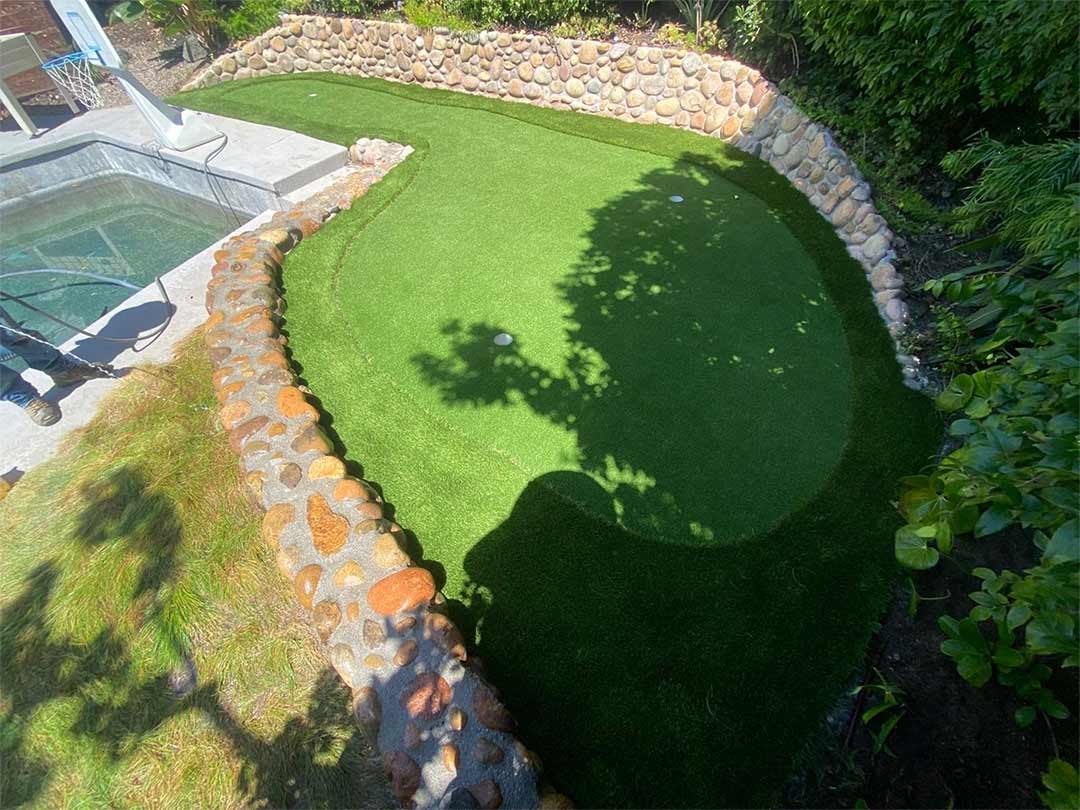 Backyard patio with gray stone tiles, a built-in grill, a rectangular fire pit filled with rocks, and artificial grass bordered by a white fence.