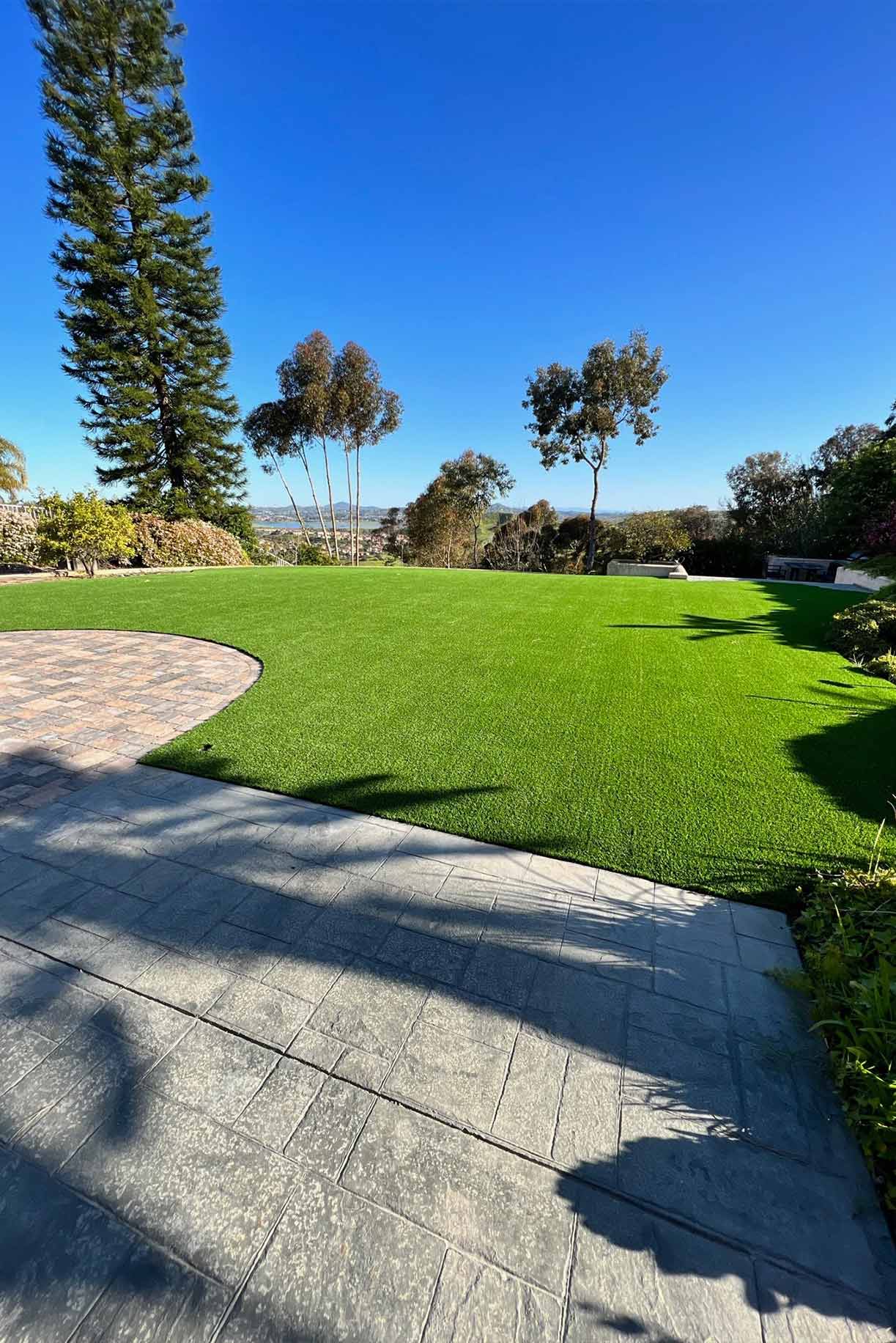 Backyard patio with a covered pergola, ceiling fans, a table holding boxes, artificial grass, and a two-story beige house.