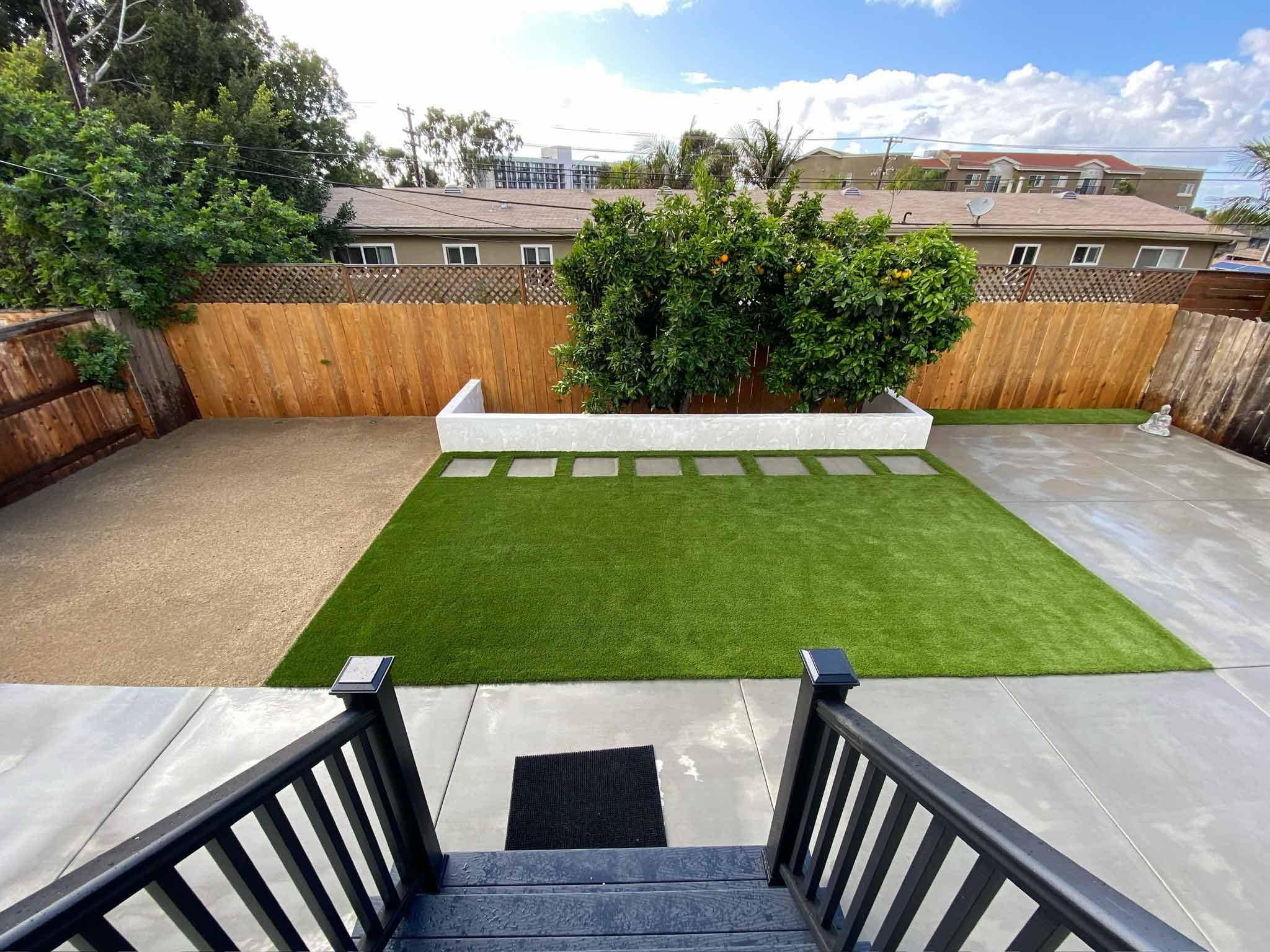 Backyard patio with a covered pergola, ceiling fans, a table holding boxes, artificial grass, and a two-story beige house.