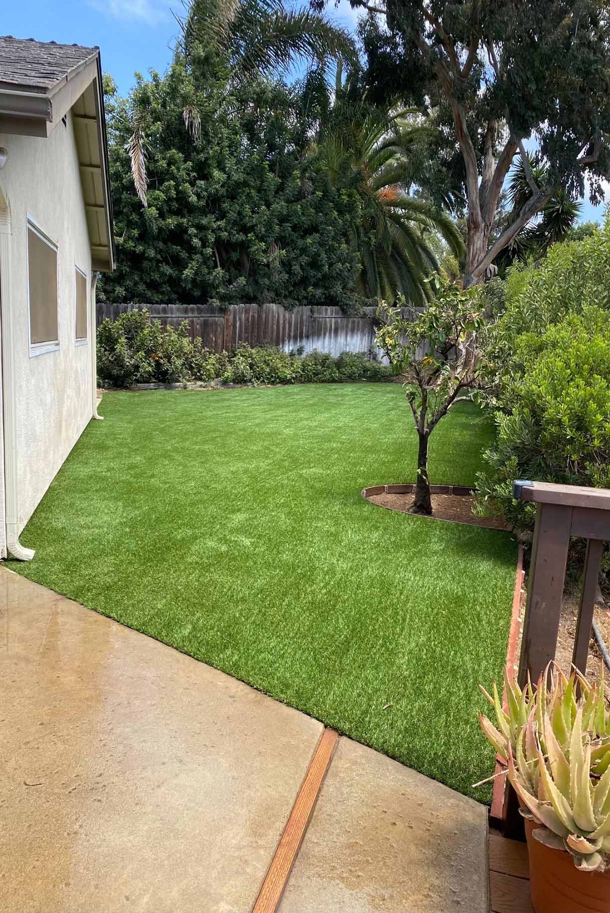 Backyard patio with a covered pergola, ceiling fans, a table holding boxes, artificial grass, and a two-story beige house.
