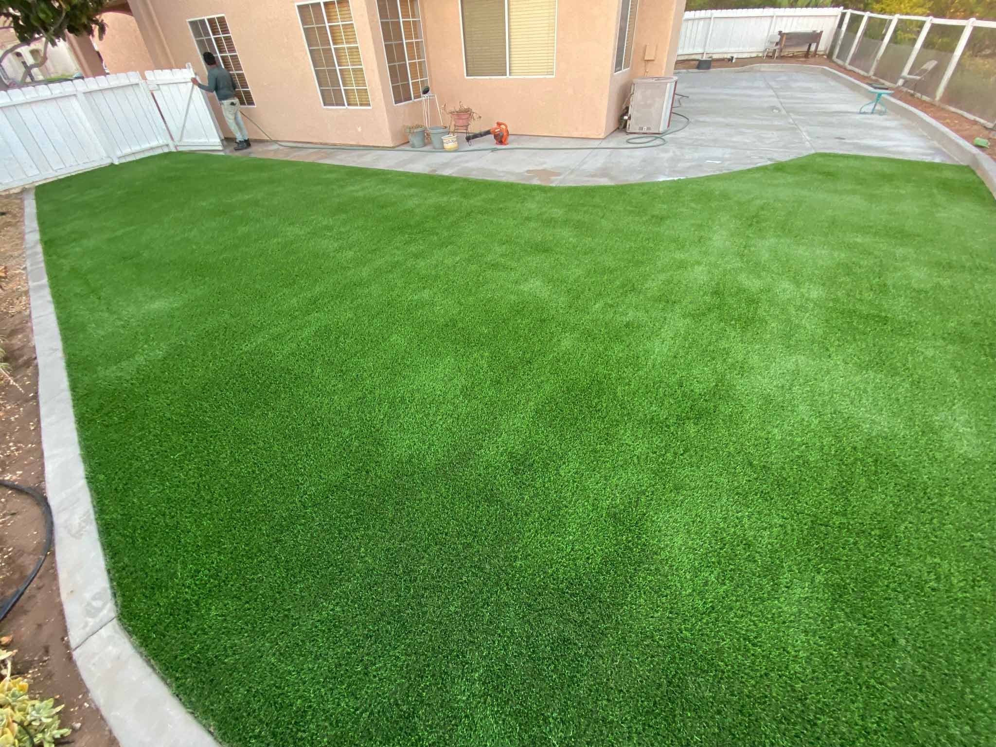 Backyard patio with gray stone tiles, a built-in grill, a rectangular fire pit filled with rocks, and artificial grass bordered by a white fence.