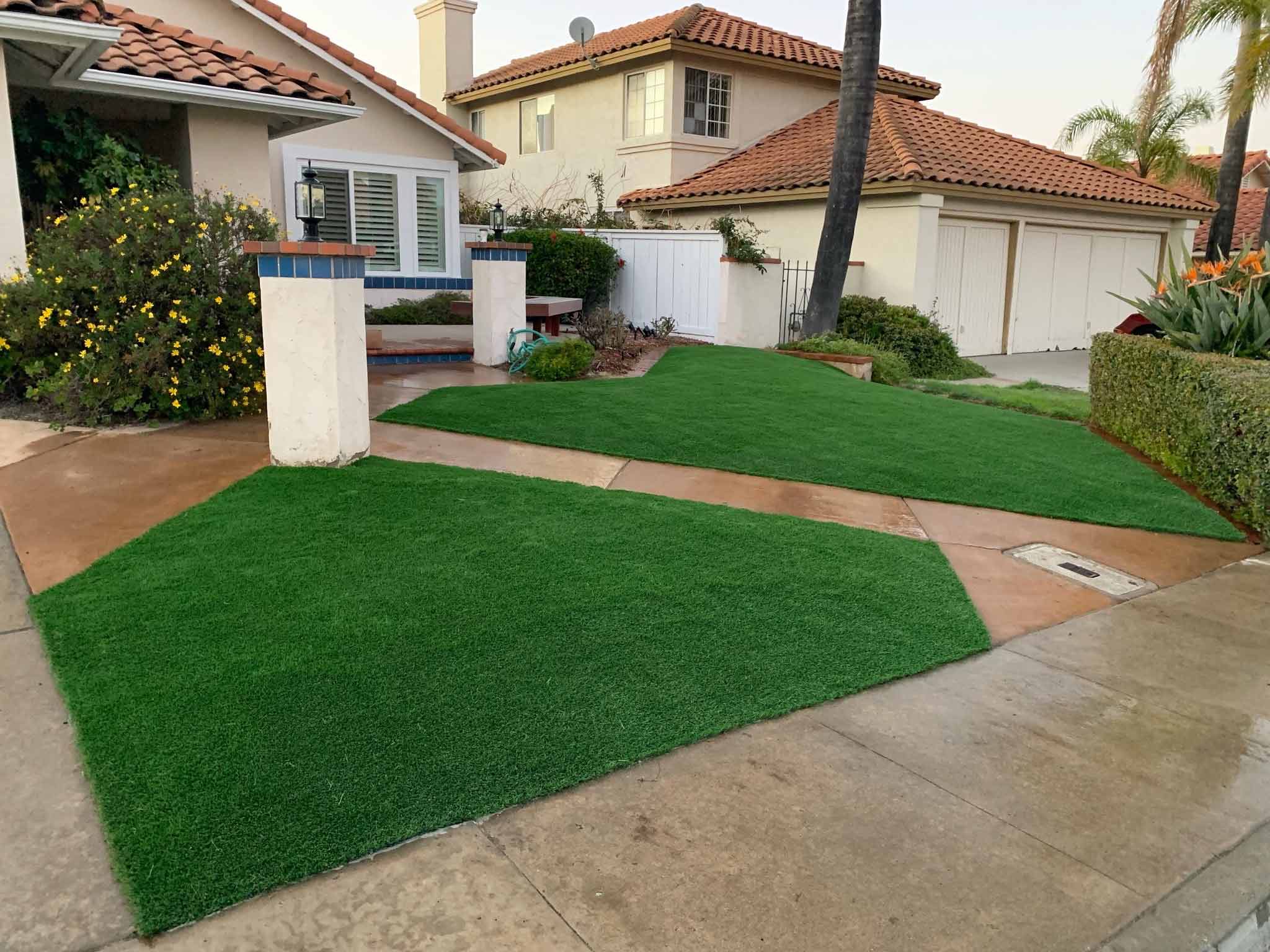 Backyard patio with gray stone tiles, a built-in grill, a rectangular fire pit filled with rocks, and artificial grass bordered by a white fence.