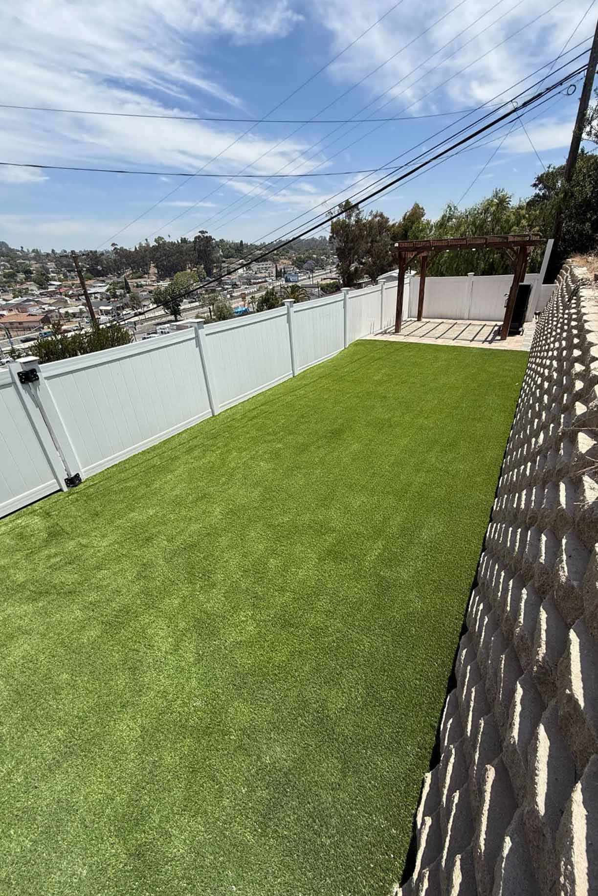 Backyard patio with gray stone tiles, a built-in grill, a rectangular fire pit filled with rocks, and artificial grass bordered by a white fence.