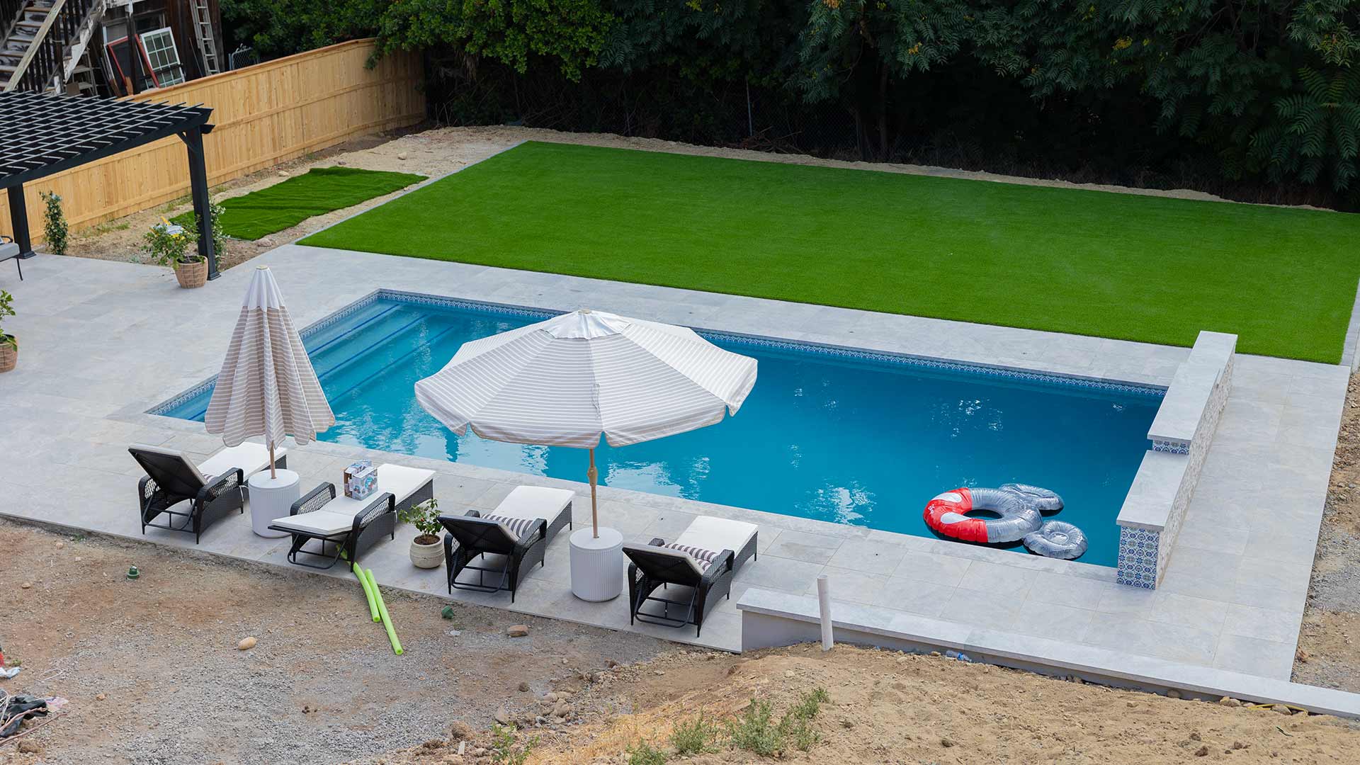 Backyard patio with a covered pergola, ceiling fans, a table holding boxes, artificial grass, and a two-story beige house.