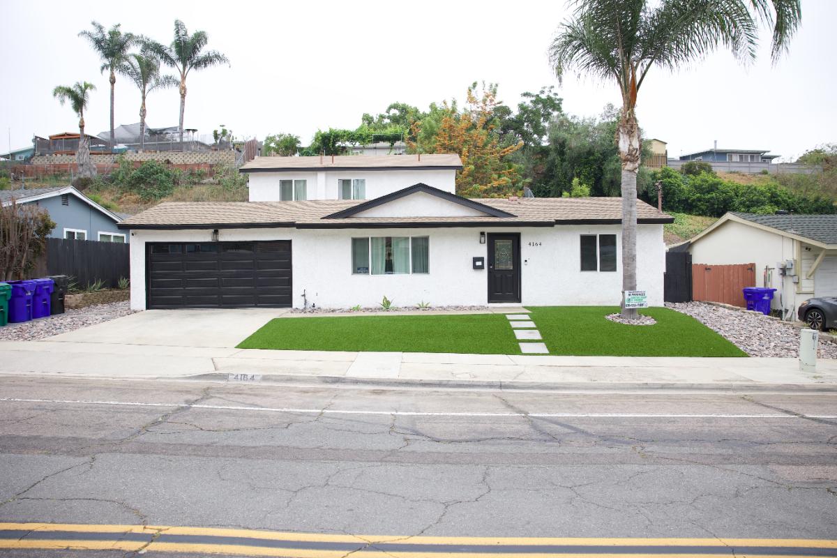 Well-manicured front yard with bright green grass, a large leafy plant cluster, a small palm tree, and a pathway leading to a blue house entrance.