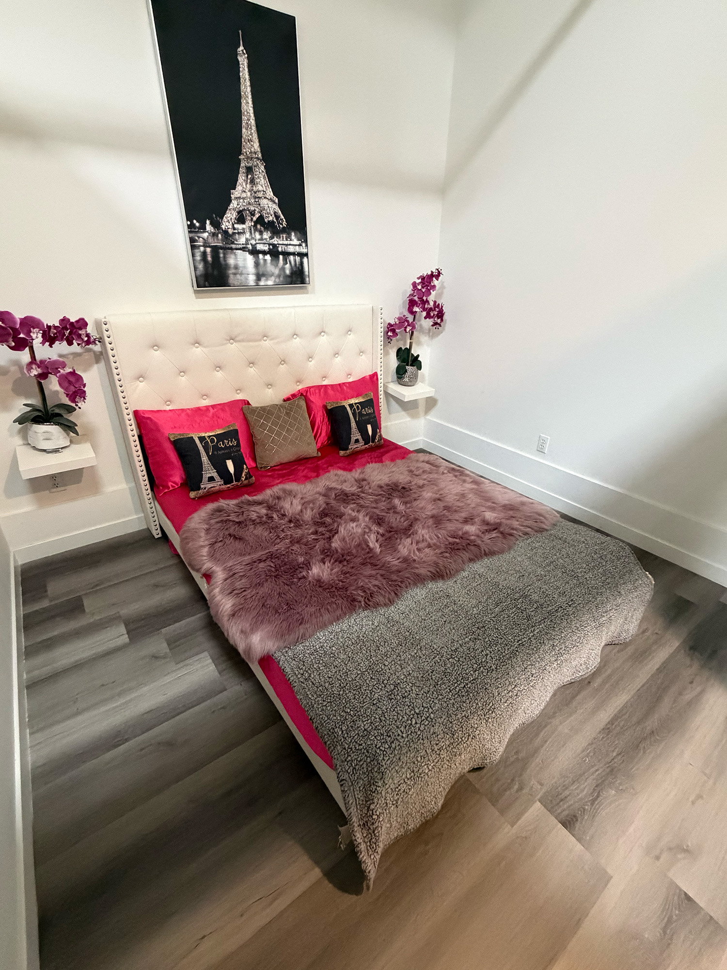 Bed with white tufted headboard, pink bedding, Paris-themed pillows, and purple flower decor on white walls.