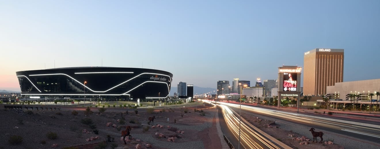Allegiant Stadium lit at dusk near busy highway with city skyline and Delano hotel in background.