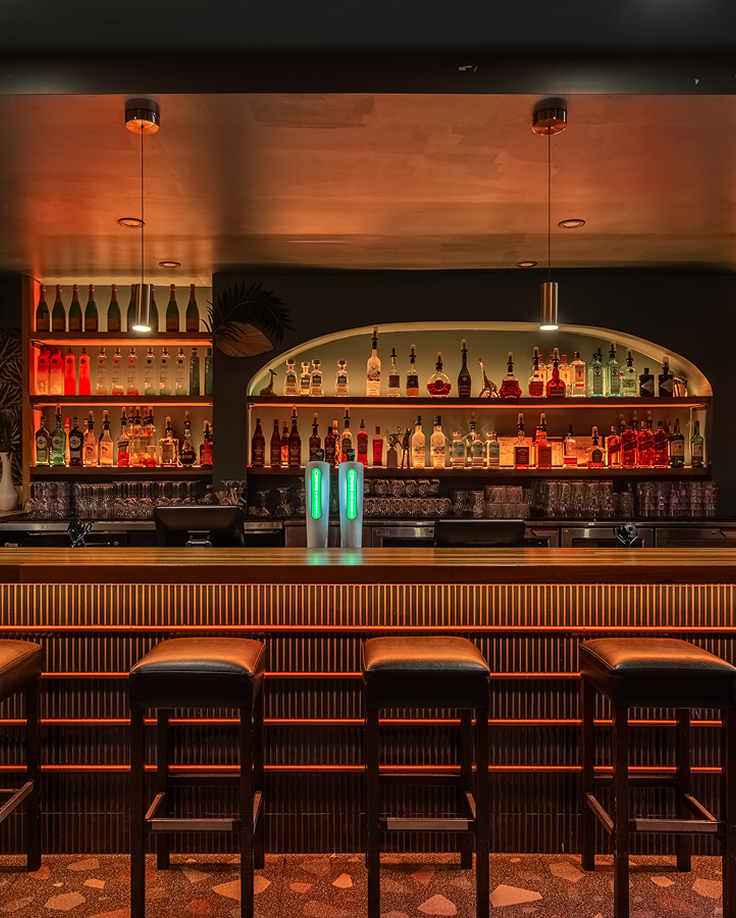 Bar moderne et éclairé avec des tabourets en cuir alignés devant une étagère remplie de bouteilles d'alcool et de verres sous un éclairage chaud.