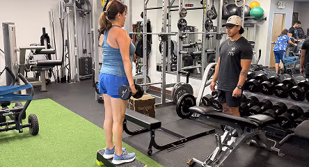 Woman lifting dumbbells on a step platform with a trainer standing nearby in a gym.