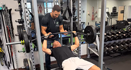 Man lying on a bench performing a barbell bench press while another man spots him in a gym.
