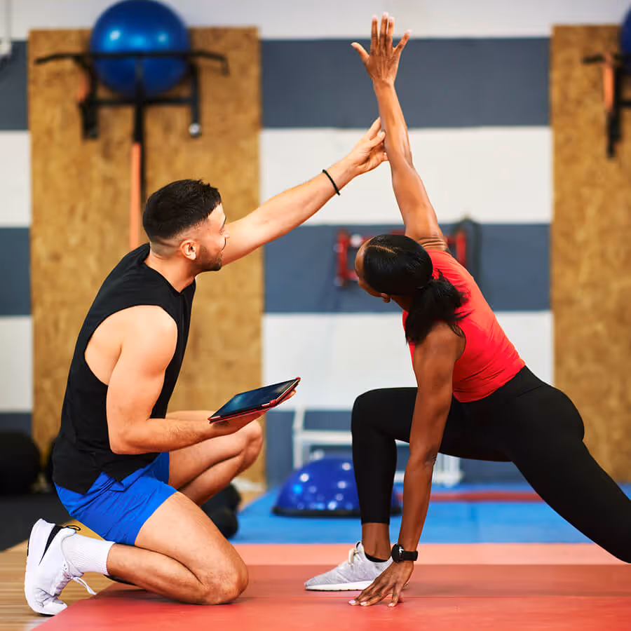 Fitness coach kneeling and assisting a woman doing a lunge stretch with one arm raised in a gym.