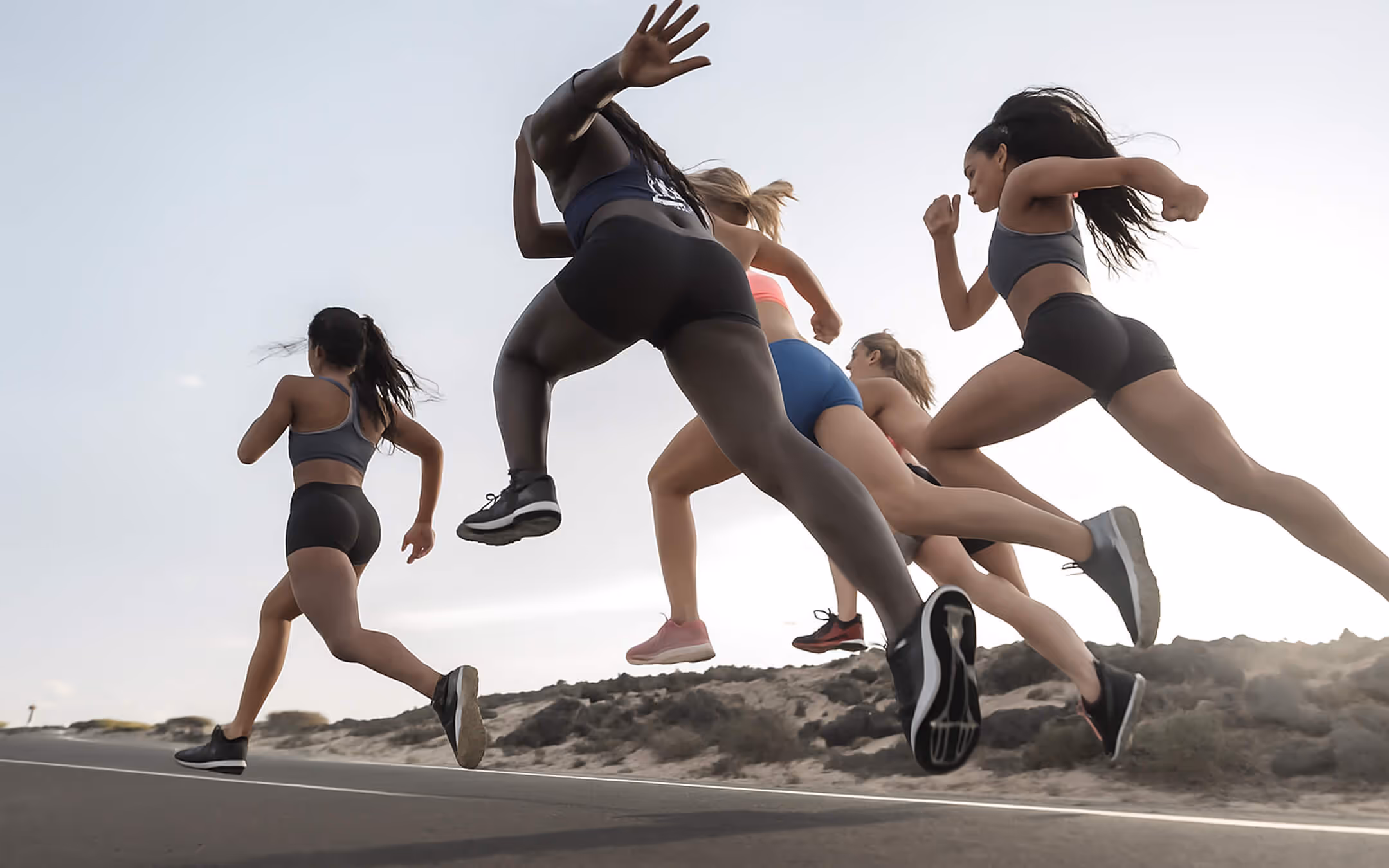 Group of women sprinting outdoors on a road with rocky terrain under a clear sky.