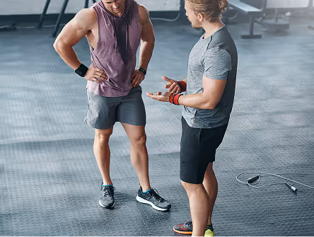 Two men in athletic clothing talking inside a gym with workout equipment in the background.