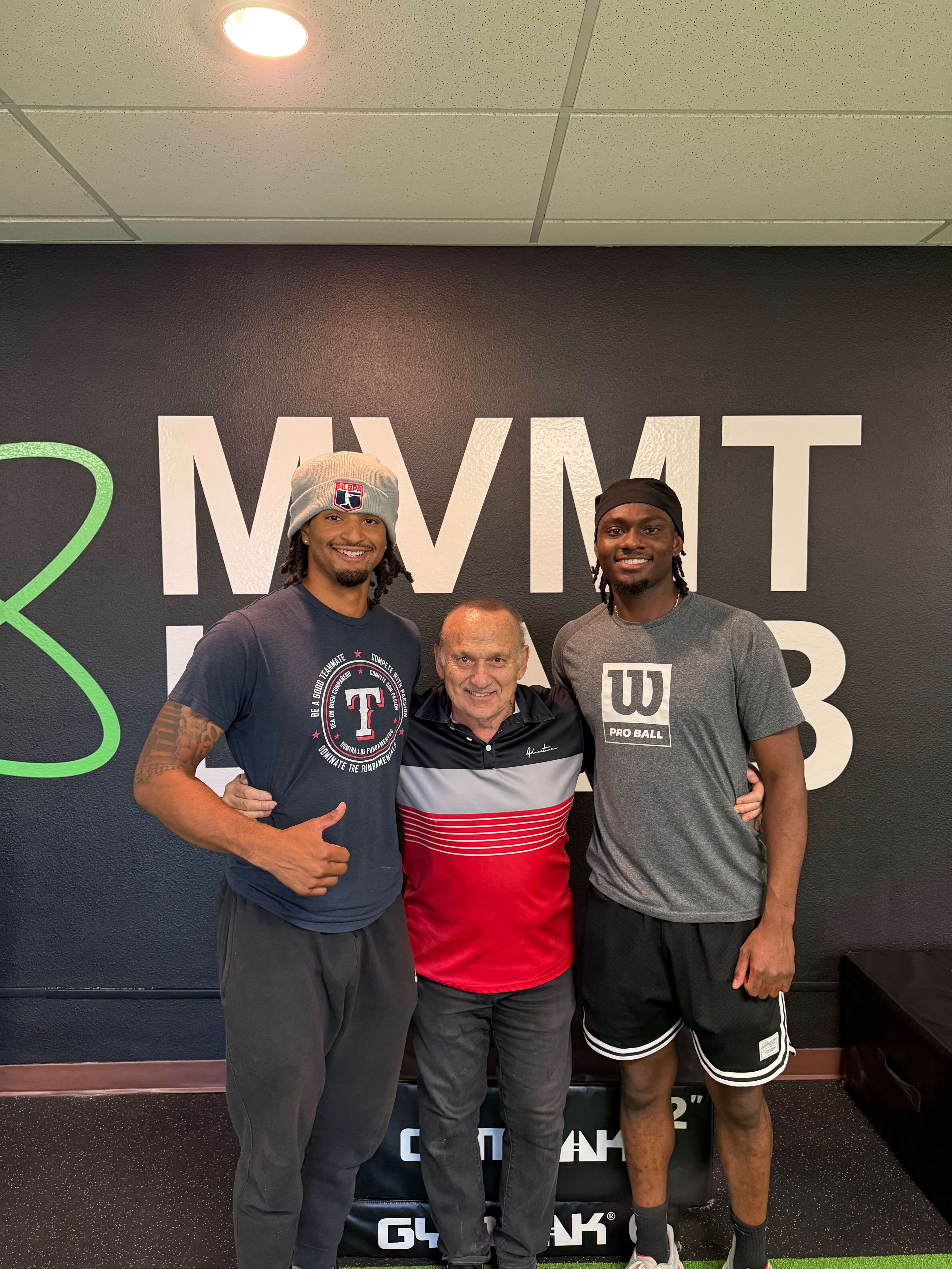 Three men smiling and posing together in a gym with a black wall background featuring large white letters and a green design.