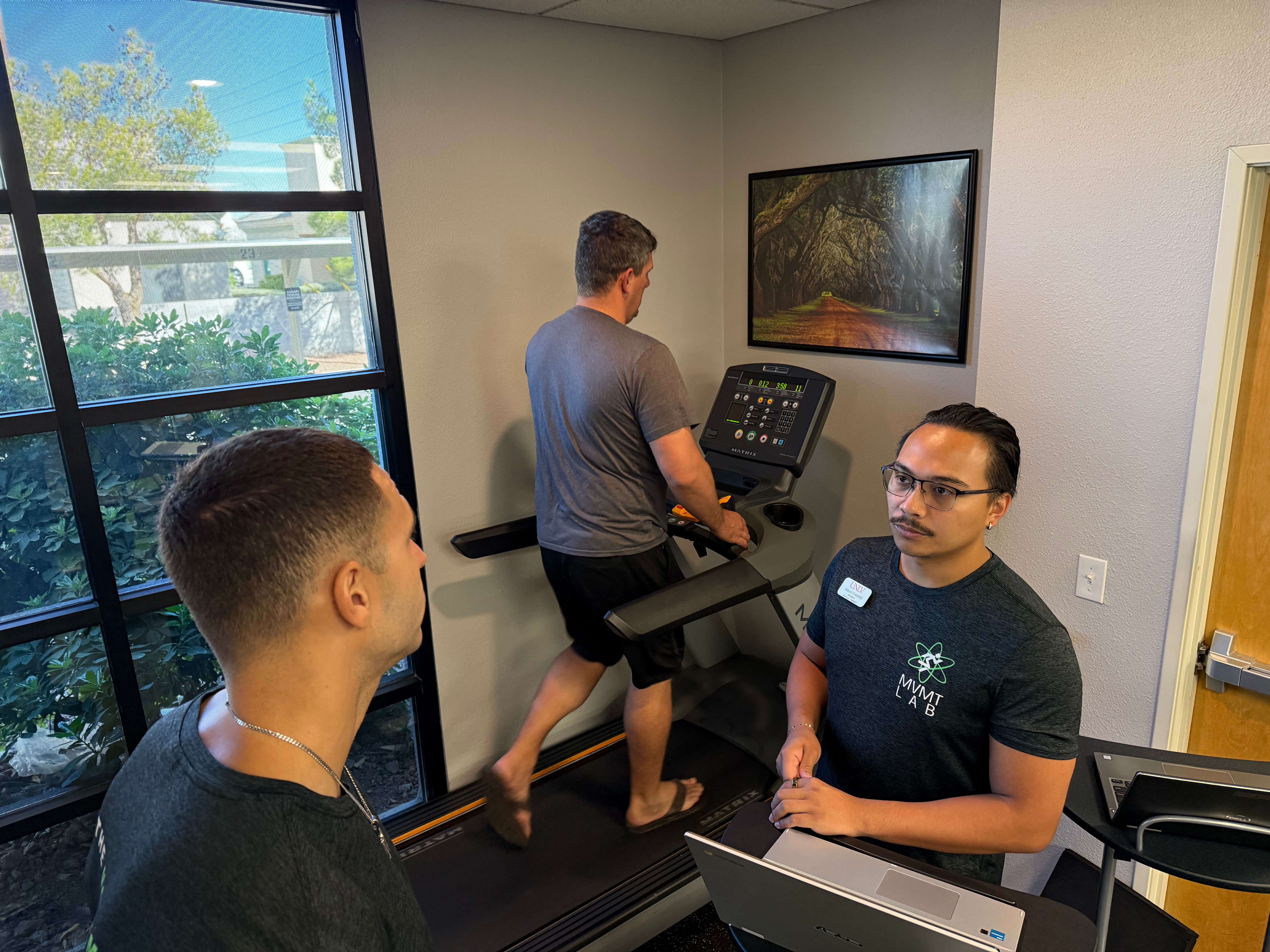 A man walking barefoot on a treadmill while another man in a MVMT Lab shirt observes and uses a laptop in a small gym room.