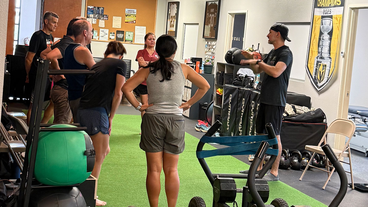 Group of people standing on green turf in a gym while a man in a black shirt and cap speaks to them.