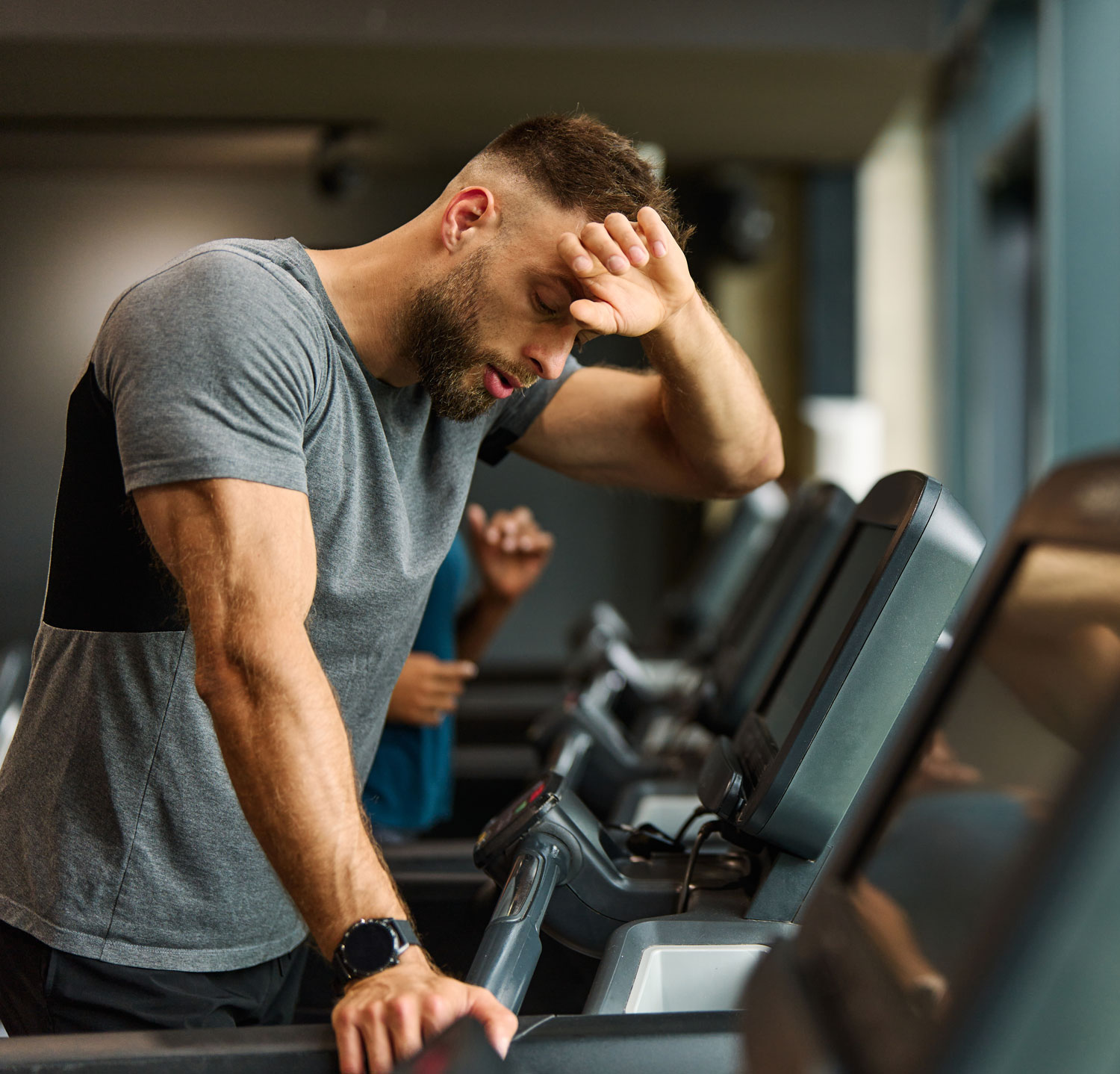 Tired man in a grey t-shirt wiping sweat from his forehead while exercising on a treadmill at the gym.
