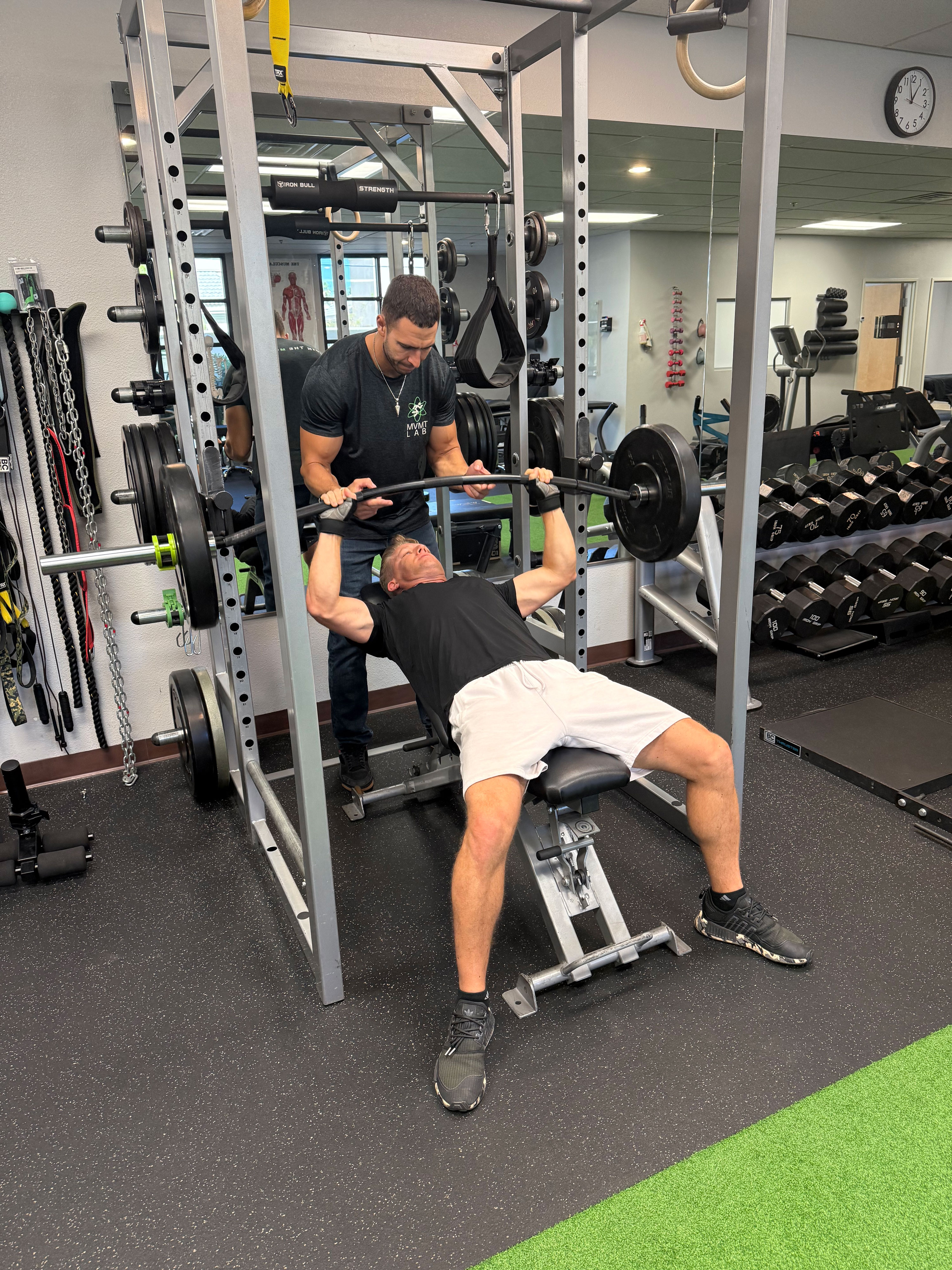Man lifting barbell on an incline bench press with a trainer spotting him in a gym.