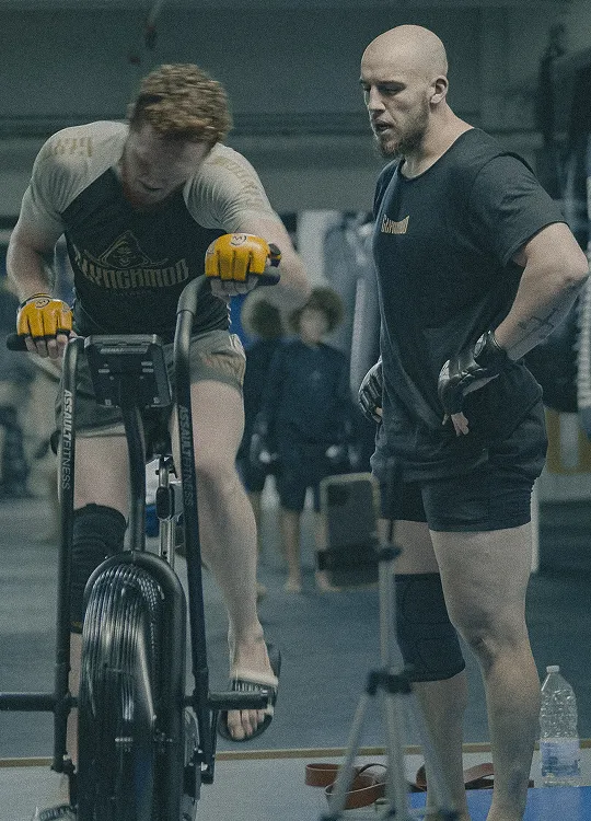 Two men in a gym, one intensely pedaling an air bike while the other stands beside him observing and coaching.