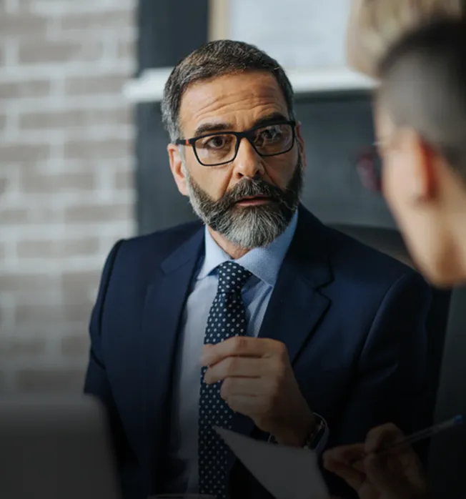 Middle-aged man with beard and glasses wearing a suit and tie, engaged in conversation with another person.