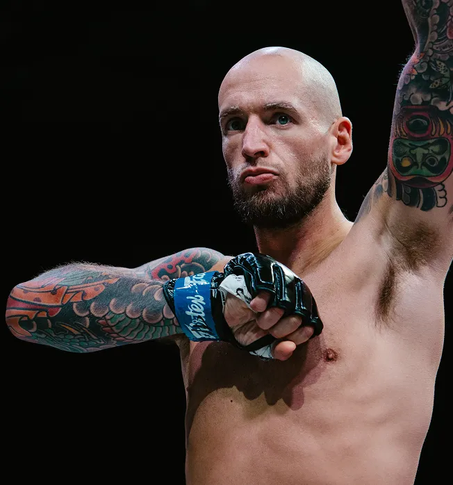 Bald tattooed male fighter with gloved hands raised in a defensive posture against a black background.