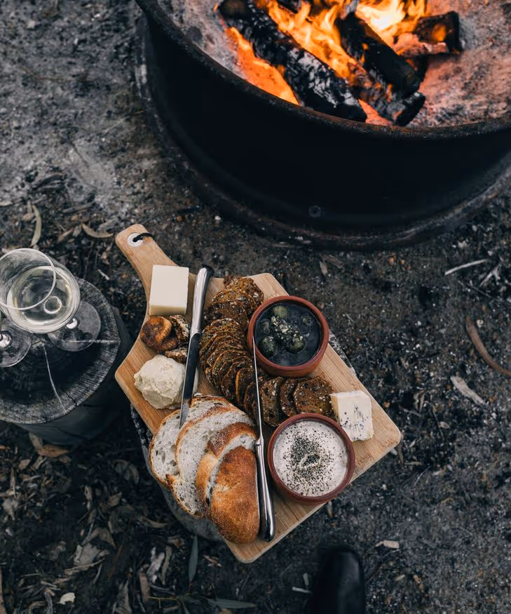 Boros plank met plakjes brood, kaas, olijven, dipsaus en crackers naast een kampvuur en twee glazen witte wijn buiten.