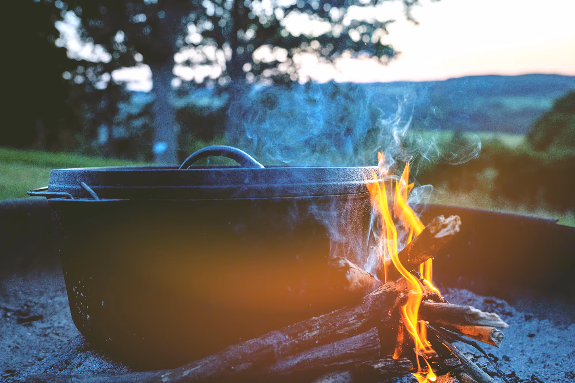 Gietijzeren pan met deksel boven een kampvuur met brandend hout en rook buiten in de natuur.