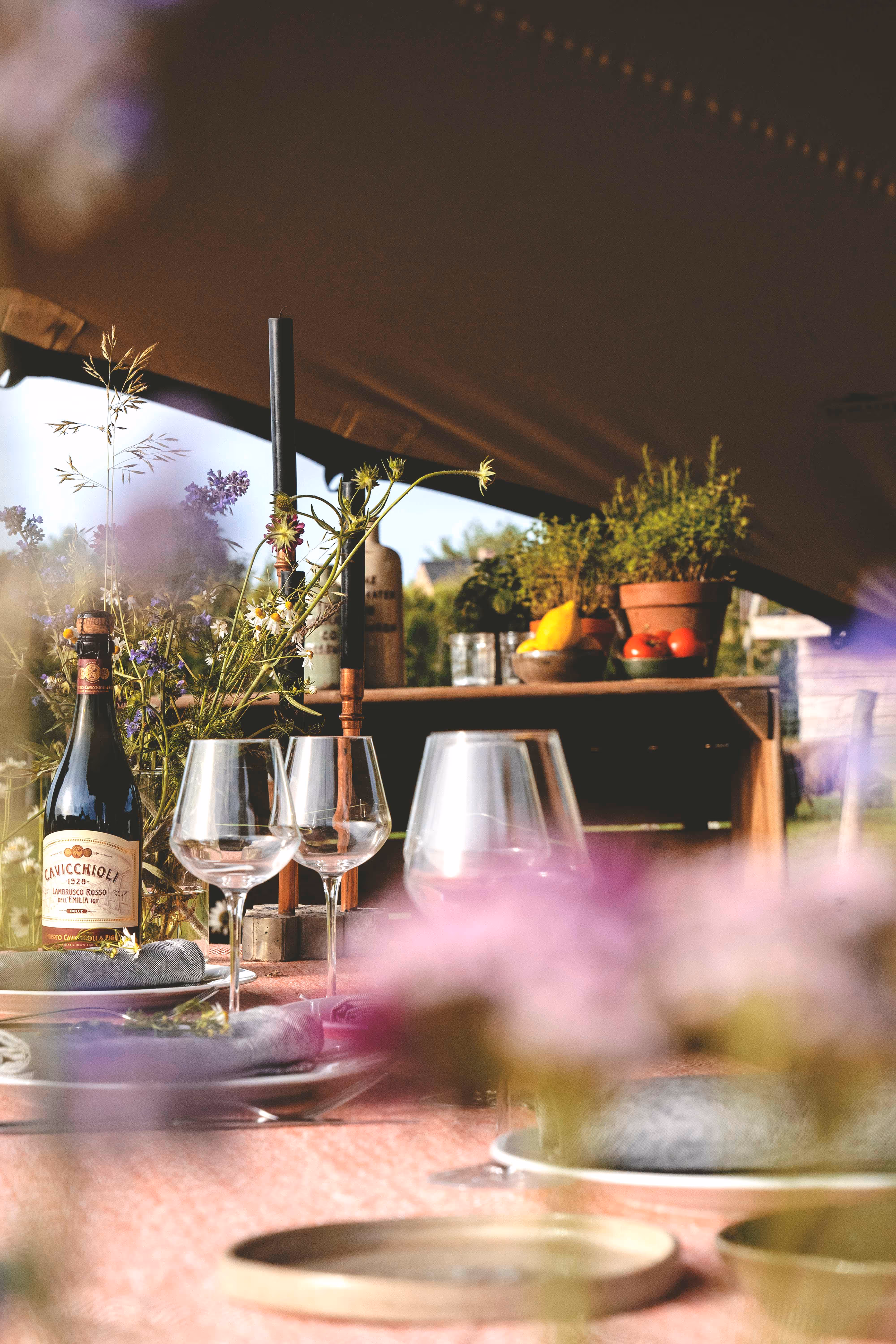 Tafeldecoratie met een fles rode wijn, lege wijnglazen, zwart kaarsen en bloemen, met planten en groentjes op de achtergrond.