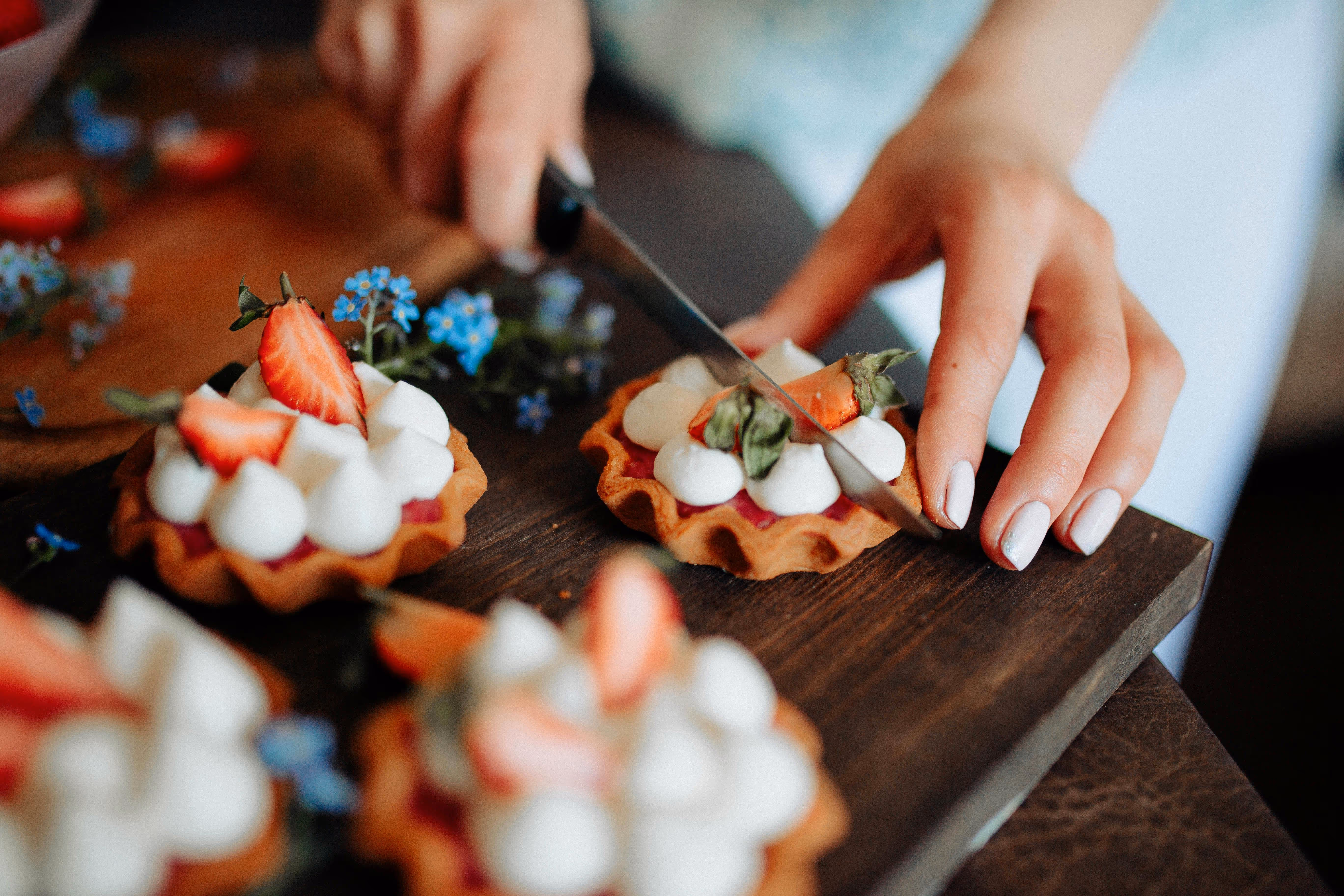 Hand met witte nagellak snijdt een kleine aardbeientaartje met room en halve aardbeien op een houten plank.
