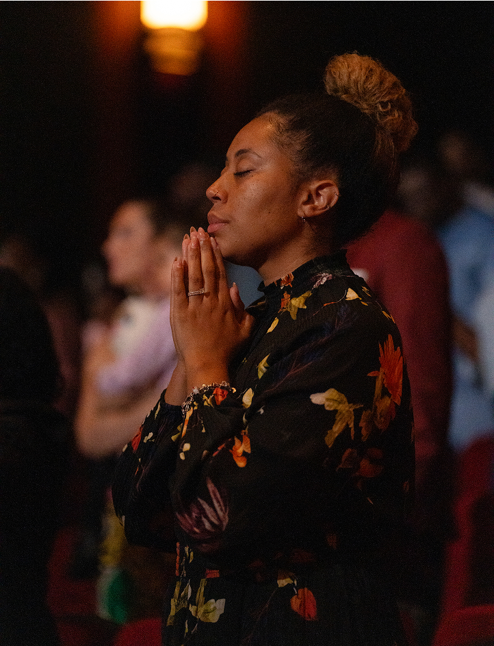 Woman with closed eyes and hands clasped in prayer wearing a floral dress in a dimly lit room.