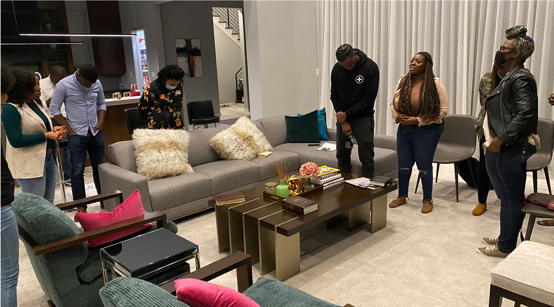 Group of people standing with bowed heads in a living room, suggesting a moment of prayer or reflection.
