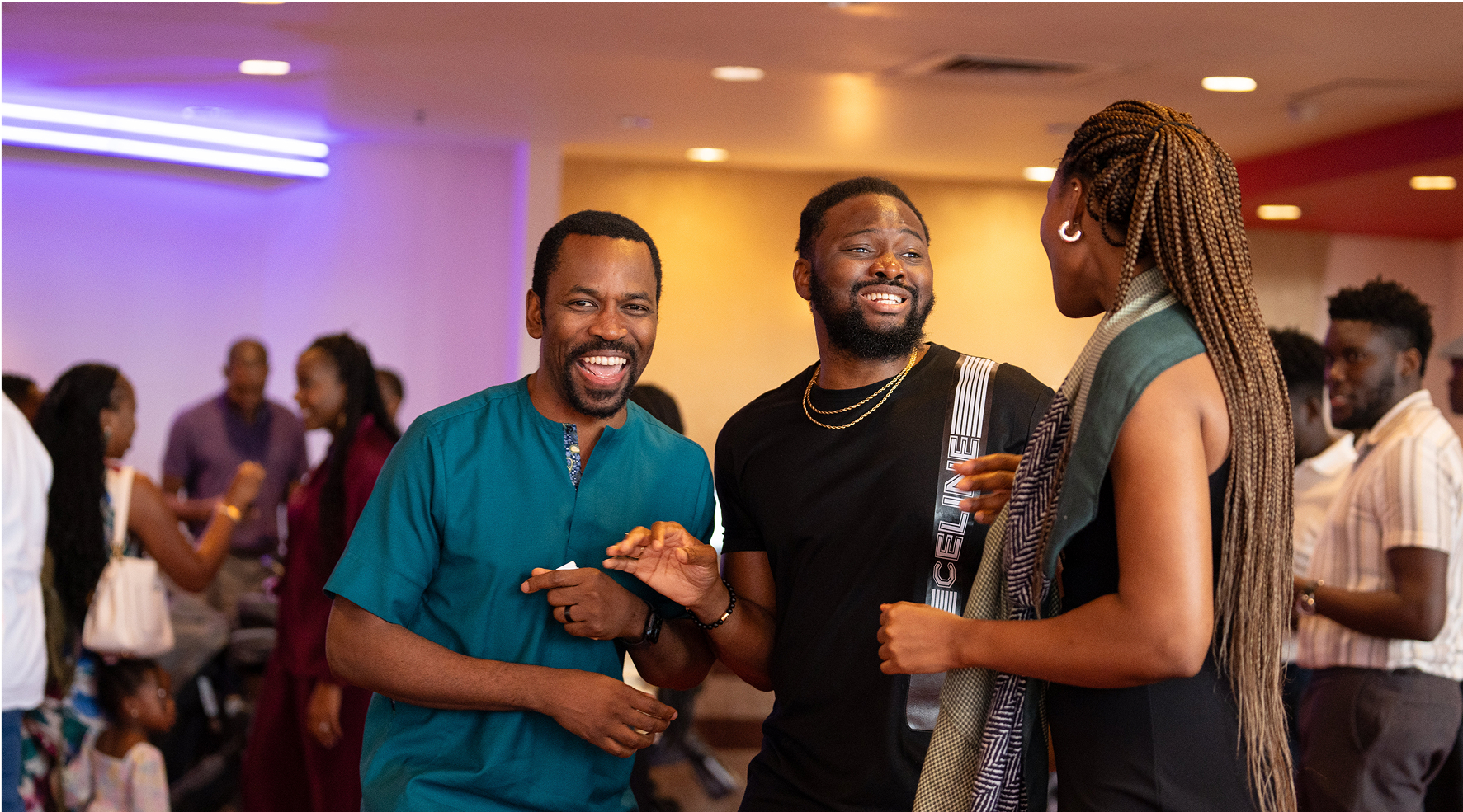 Group of people socializing indoors, two men and a woman in the foreground smiling and talking.
