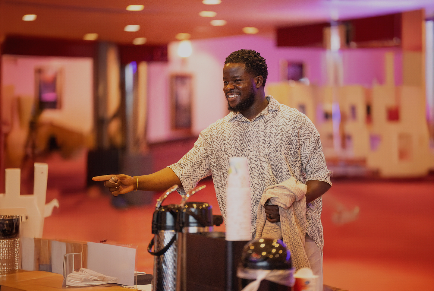 Smiling man pointing and holding a beige sweater near a coffee station in a warmly lit indoor setting.