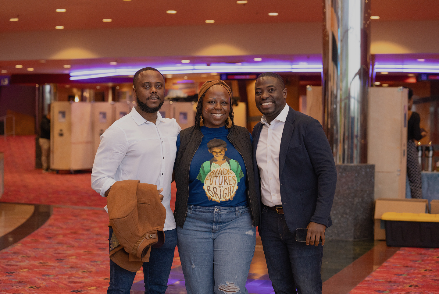Three smiling people standing together indoors on a red carpeted floor with one person wearing a blue shirt that says Black Futures Are Bright.