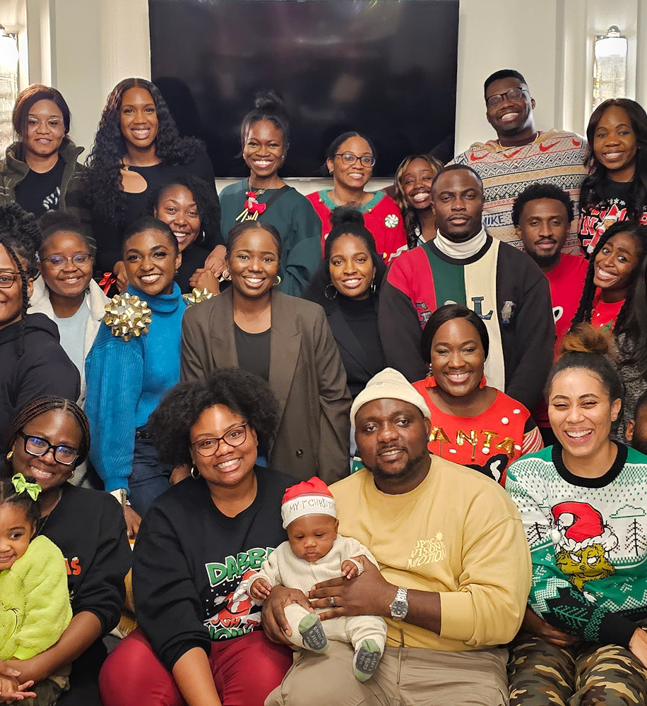 Group portrait of diverse smiling adults and children wearing festive sweaters and holiday attire indoors.