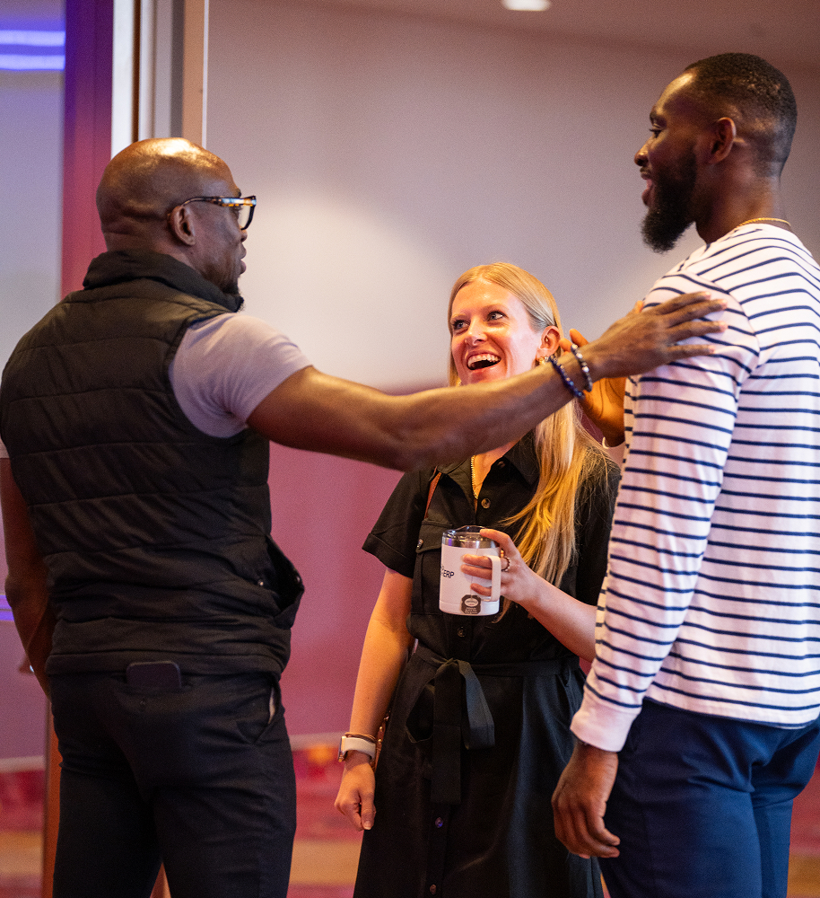 Three diverse adults smiling and conversing indoors, one man wearing glasses and a black vest gestures while a woman holding a mug listens.