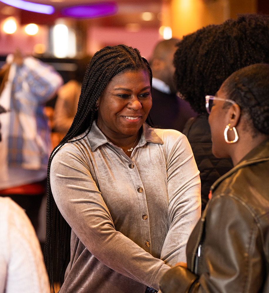 Smiling woman with braided hair shakes hands with another person in a warm, indoor social setting.