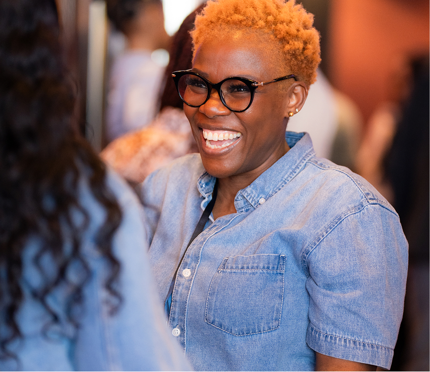 Smiling woman with short orange hair and glasses wearing a denim shirt, engaging in conversation.