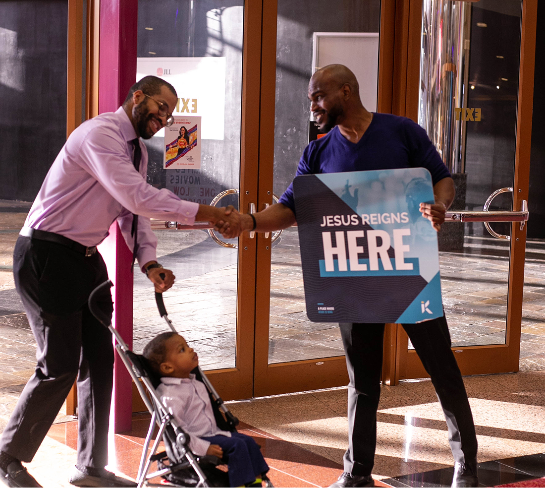 Two men shaking hands near glass doors; one is holding a sign that reads 'Jesus reigns here,' and a child sits in a stroller nearby.