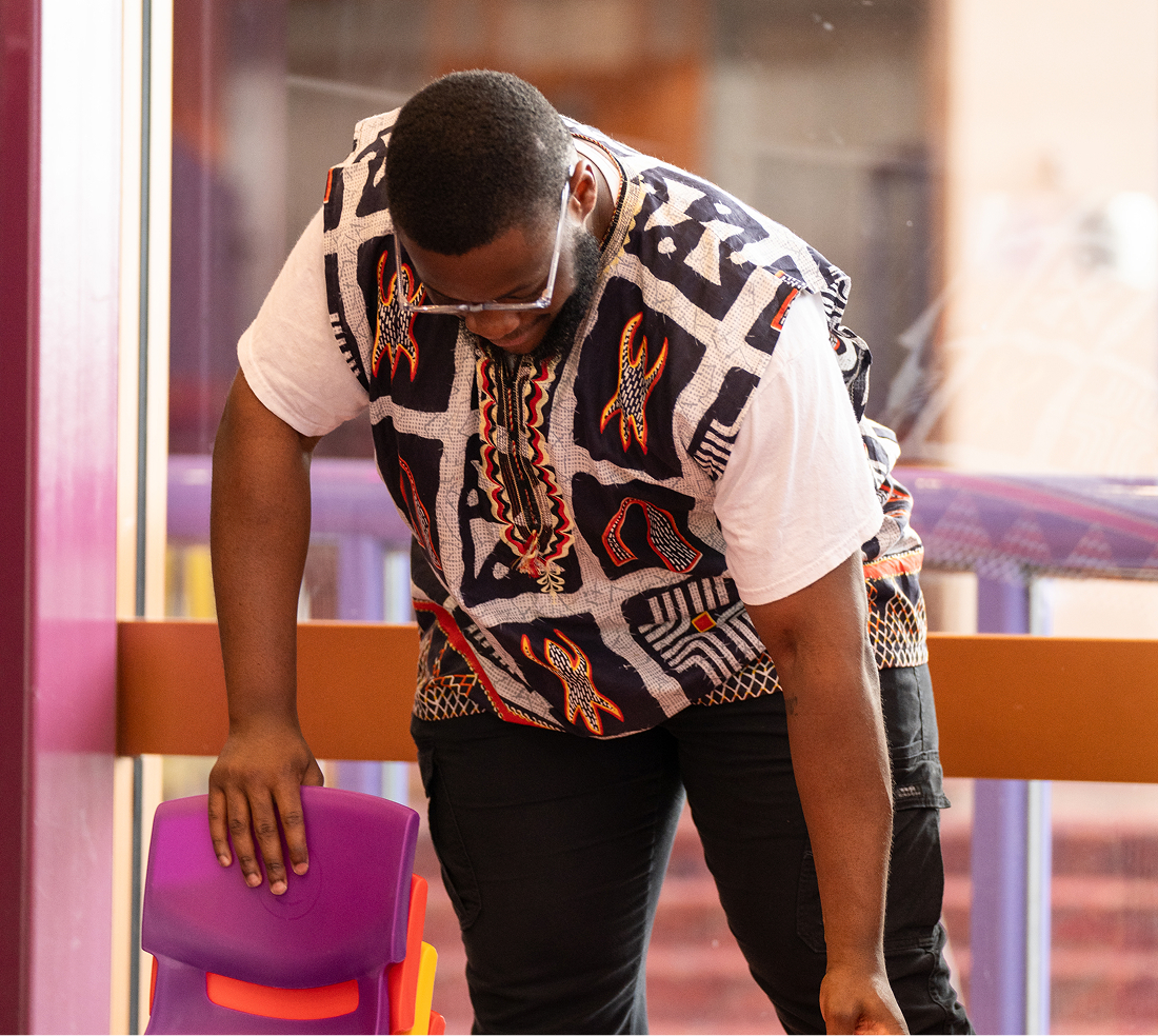 Man wearing glasses and a patterned shirt bending over while touching a stack of colorful plastic chairs.