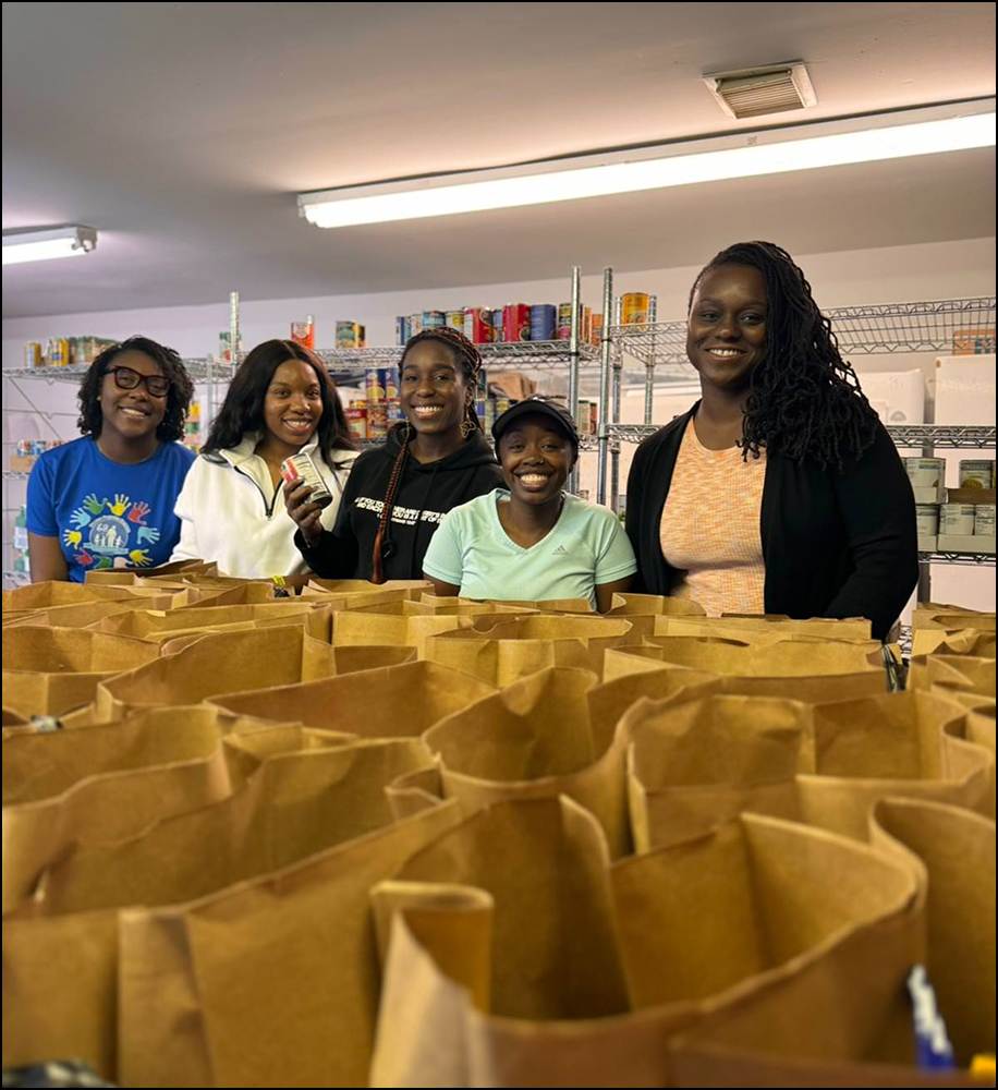 Five smiling women standing behind rows of brown paper bags in a food pantry with shelves of canned goods in the background.