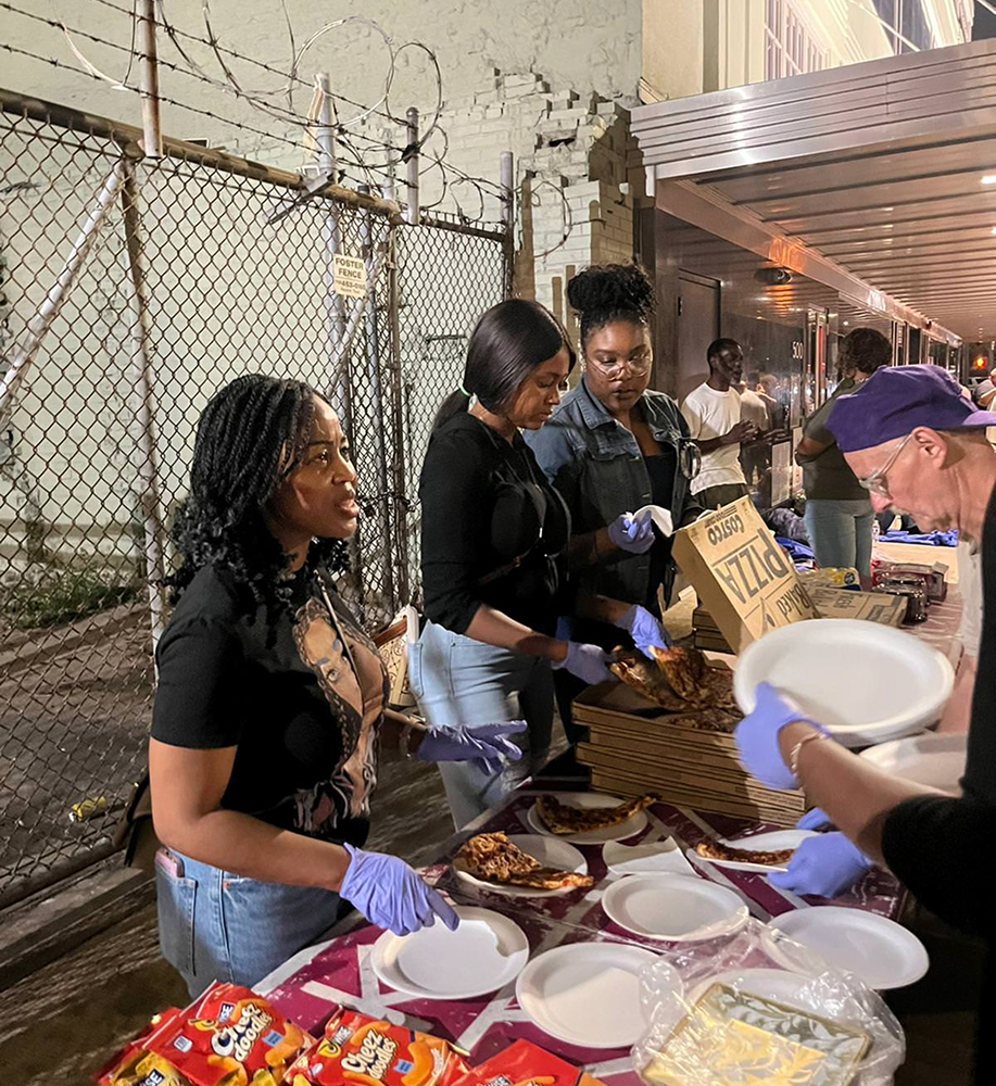 Three women wearing gloves serve pizza slices to people at an outdoor table with snacks and paper plates at night.