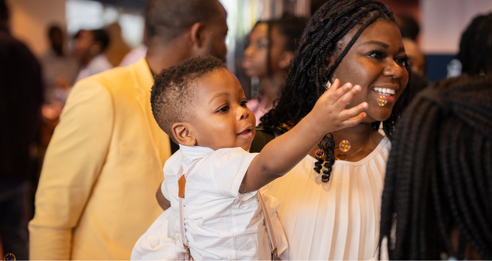 Smiling woman holding a toddler boy who is reaching out with his hand in a lively indoor setting.