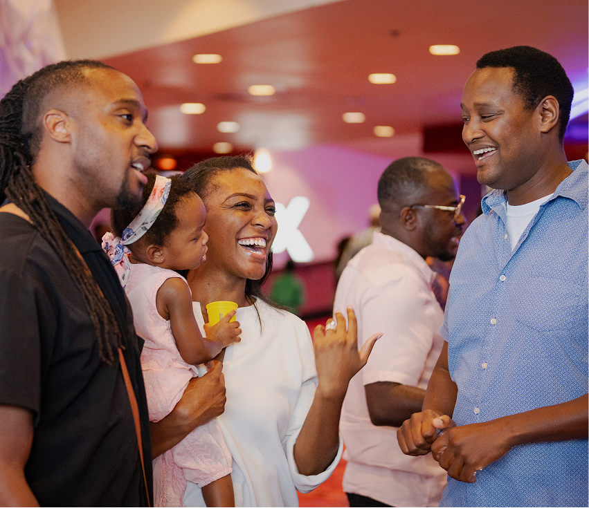 A smiling woman holding a toddler talks with two men in a brightly lit indoor setting.